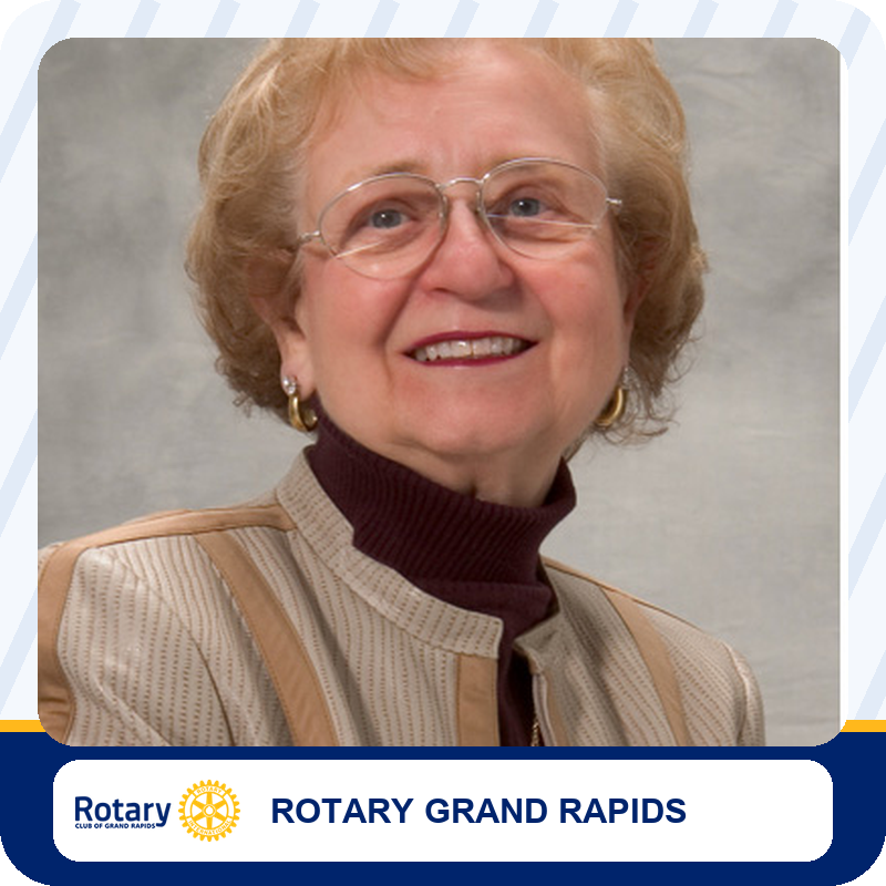 Smiling elderly woman with short blonde hair and glasses, wearing a beige jacket over a dark maroon turtleneck.