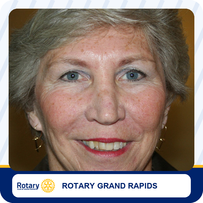 Close-up portrait of a smiling older woman with short gray hair and blue eyes on a Rotary Grand Rapids banner.