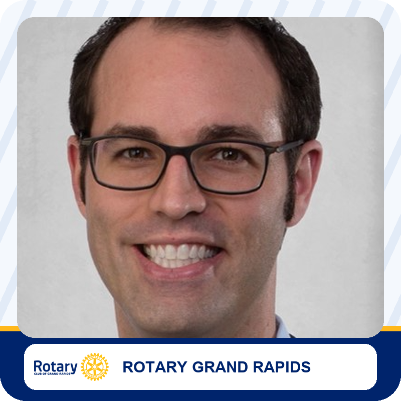 Smiling man with short dark hair and rectangular glasses in a head-and-shoulders portrait against a gray backdrop; Rotary Grand Rapids branding appears below.