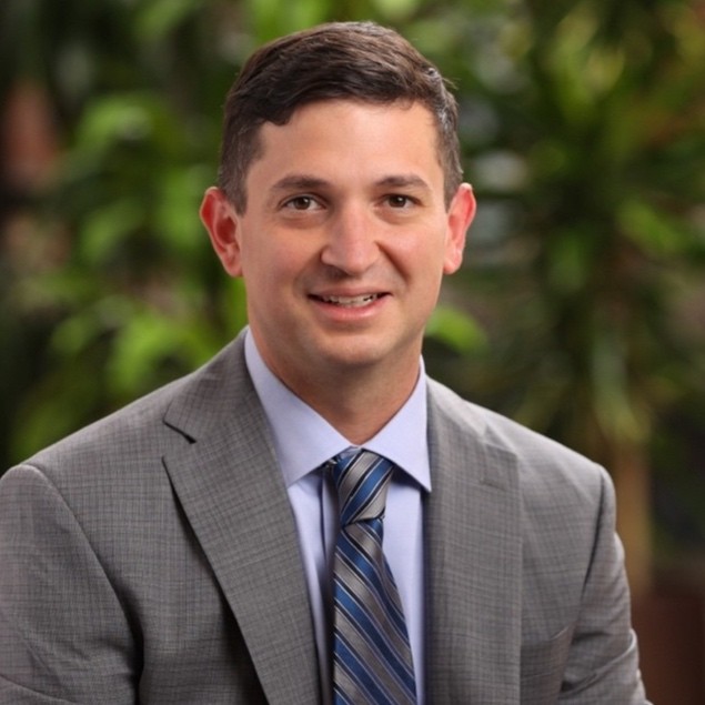 Professional headshot: man in a gray suit and blue striped tie, smiling, with greenery in the background.