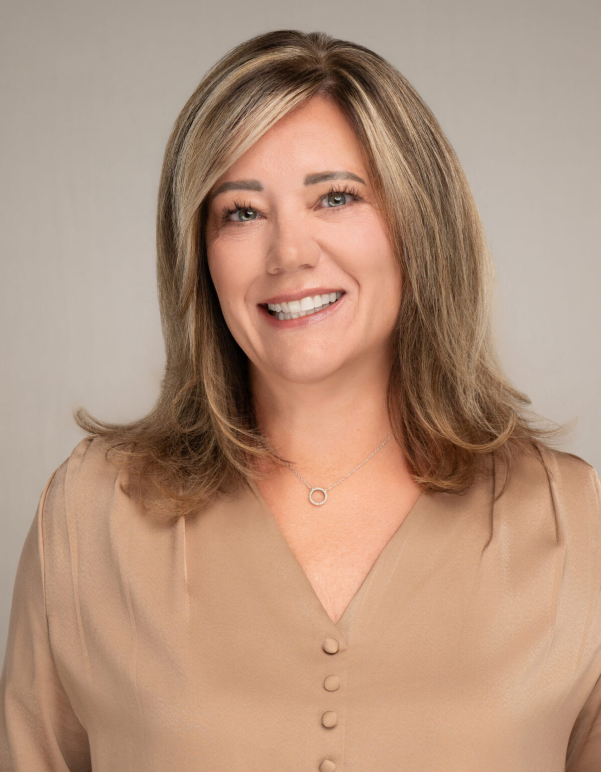 Smiling woman with shoulder-length blonde-highlighted hair in a beige blouse and silver circular pendant necklace against a neutral backdrop.