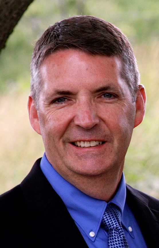 Smiling man in a blue dress shirt and patterned tie, outdoors with a blurred natural background.